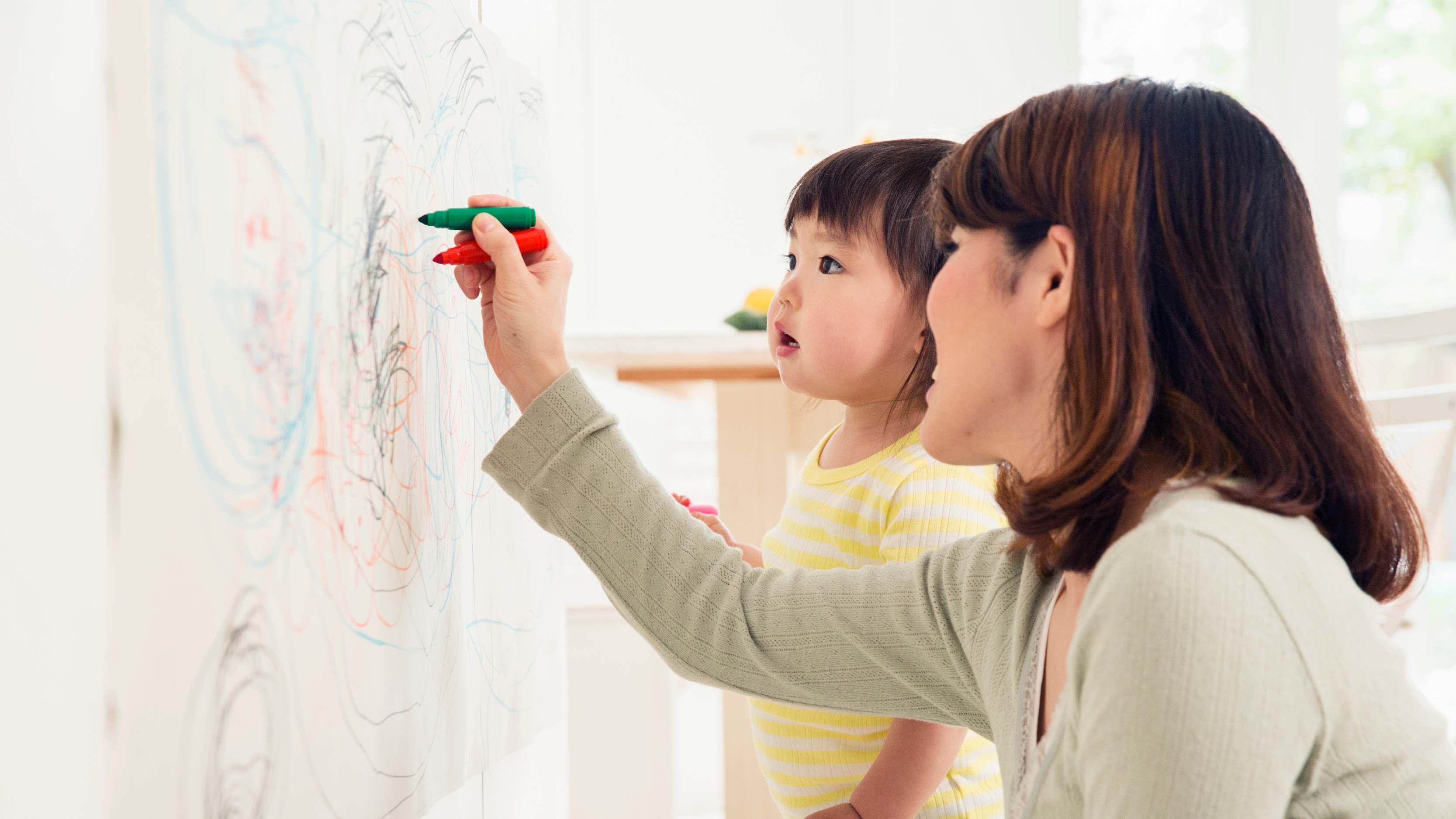 A woman and a child stand in front of a large board. The woman is writing on it.