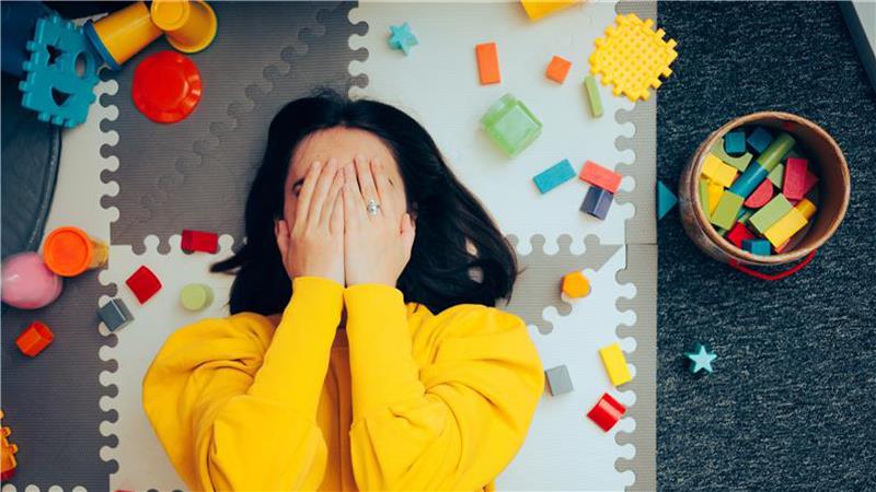 An image of a female caregiver lying face-up on the floor surrounded by toys, holding her hands over her face in exhaustion.